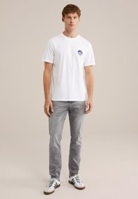 Camiseta de algodón blanca de manga corta, con un gráfico azul en el lado izquierdo del pecho. Llevada con jeans gris claro y zapatillas blancas.