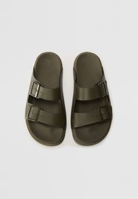 Olīvzaļas slīdošās sandaljas, izgatavotas no tekstruētā sintētiskā materiāla, ar divām regulējamām siksnām un sprādzēm drošai piegulšanai.