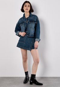 Denim-Jacke und Rock im Dunkelblau mit hellblauen Akzenten; verfügt über einen spitzen Kragen, eine Knopfleiste und aufgesetzte Taschen. Kombiniert mit schwarzen Stiefeletten.