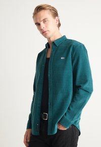 Camisa de pana color teal con cuello de botón, que presenta un pequeño logo en el lado izquierdo del pecho, llevada sobre una camiseta sin mangas negra y un cinturón negro.