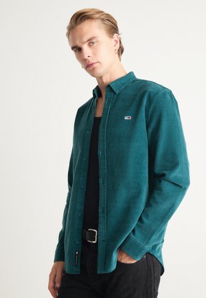 Camicia in velluto a coste color teal con colletto a bottoni, dotata di un piccolo logo sul lato sinistro del petto, indossata sopra un top nero senza maniche e una cintura nera.