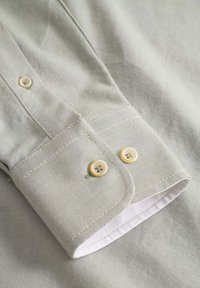 Helles graues Hemd mit einem Knopfmanschette, ausgestattet mit zwei cremefarbenen Knöpfen und kontrastierendem weißen Innenfutter. Glattes Gewebe mit feiner Textur.
