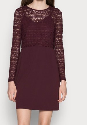 Frau trägt ein burgunderrotes Kleid mit spitzenbesetzten langen Ärmeln und Spitzenüberzug am Oberteil, einfarbiger Rock, steht vor einfachem Hintergrund.