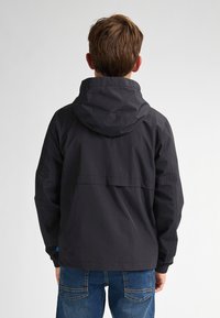 Jachetă ușoară neagră cu glugă, având o croială lejeră, manșete elastice și un tiv drept. Purtați peste blugi albaștri, afișată din spate.