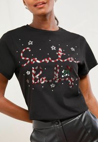Čierne tričko s nápisom "Santa Baby" v červenej a bielej sequínovej literi, zdobené striebornými hviezdami a zelenými detailmi imela.