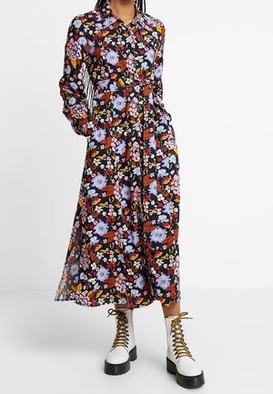 Vrouw draagt een lange mouw, gebuttonde maxi-jurk met bloemenprint, een kraag en witte vetercombatlaarzen met dikke zwarte zolen.