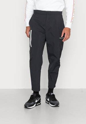 Homme portant un pantalon noir fuselé avec des poches latérales zippées, des baskets Nike noires et une chemise blanche à manches longues avec des rayures rouges sur les manches.
