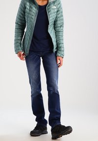 Nafouklá tyrkysová bunda s prošívanou texturou, tmavě modré tričko, modré džíny a černé trekové boty se žlutými detaily. Osoba stojící čelně.