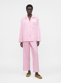 Γυναίκα που φορά χαλαρό ροζ και λευκό πουκαμισοειδές pyjama με λεπτές ρίγες και παντελόνι με λευκή λουλουδένια λεπτομέρεια στην τσέπη στο στήθος, συνοδευόμενο από καφέ υφαντά φλατ παπούτσια.