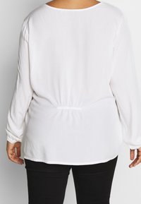 White long-sleeve blouse made of lightweight fabric, featuring a round neckline, elastic waist, and gathered shoulder detail.