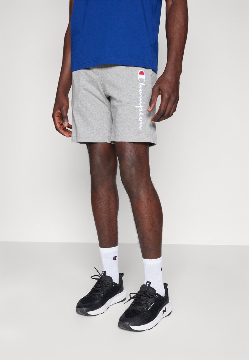 Shorts de algodón grises con el logo blanco de "Champion" en el lado, combinados con zapatillas deportivas negras y calcetines blancos con logo, llevados con una camiseta azul.