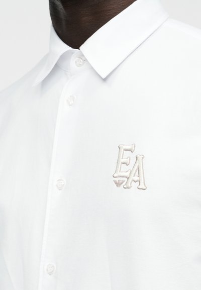 Άνδρας που φοράει λευκό πουκάμισο με κουμπιά, με μυτερό γιακά και κεντημένα αρχικά "EA" στο στήθος.