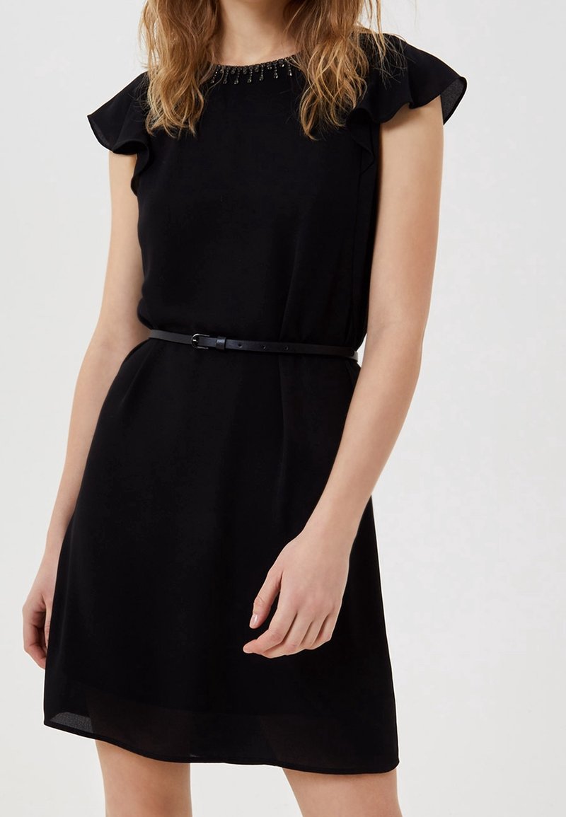 Femme portant une robe noire sans manches avec des manches à volants, un col orné de perles et une fine ceinture noire à la taille, debout devant un fond uni.