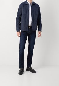 Giacca blu navy con chiusura a bottoni e quattro tasche frontali, indossata sopra una camicia bianca e jeans scuri, abbinata a mocassini neri.