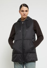 Kvinna i svart ärmlös pufferväst med huva över en brun långärmad tröja, med händerna i västfickorna, mot en enfärgad ljus bakgrund.