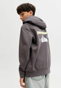 Ragazzo che indossa un felpa grigio scuro con testo giallo e una grafica sul retro, abbinato a jeans di denim blu chiaro, in piedi contro uno sfondo semplice.