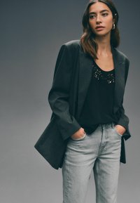 Donker gestreepte blazer over een zwart tanktop met kantdetail, gecombineerd met lichtgrijze spijkerbroek. Soepele stoffen en een relaxte pasvorm.