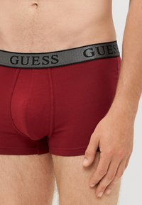 Slip in boxer rossi con una cintura elastica nera che presenta il logo "GUESS" in lettere argentate. Il materiale sembra morbido e aderente.