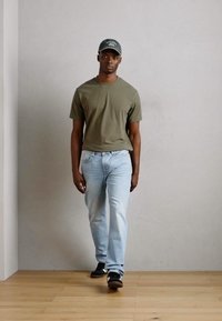 Olive green t-shirt, light blue denim jeans, black sneakers, and a dark cap worn by a person walking in a minimalistic indoor setting.