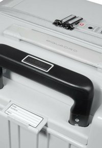 Maleta gris con ruedas y asa negra. Cuenta con una cremallera blanca, detalles metálicos y un portaetiquetas para nombre/dirección. Diseño compacto y duradero.