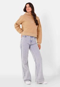 Sudadera con capucha beige de diseño cropped y con dobladillo acanalado, combinada con jeans acampanados de color azul claro y zapatillas blancas. Textura suave, estilo casual.