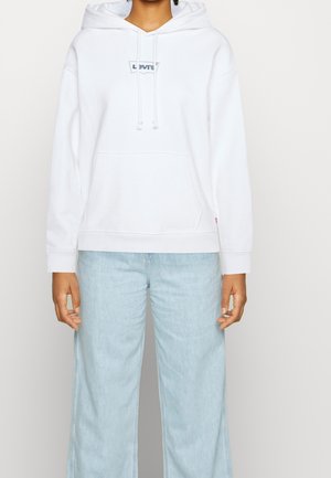 Osoba nosząca biały kaptur Levi’s z przednią kieszenią i sznurkami, w połączeniu z jasnoniebieskimi szerokimi dżinsami.