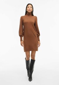 Donna che indossa un vestito marrone a maniche lunghe con colletto alto e stivali neri fino al ginocchio, in piedi contro uno sfondo bianco.