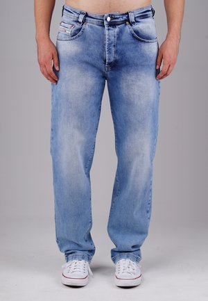 Man wearing light blue faded jeans and white sneakers standing against a plain gray background, showing lower torso and legs.