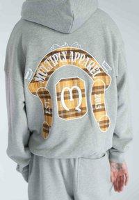 Šedá mikina s kapucňou s veľkým grafickým motívom na chrbte s textom "MULTIPLY APPAREL" a kockovaným vzorom v žltej a hnedej farbe.