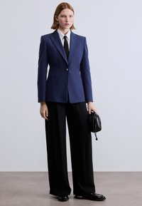 Σκουρόχρωμο μπλέιζερ ναυτικής έμπνευσης με δομημένους ώμους, κλείσιμο με ένα κουμπί και δύο τσέπες, σε συνδυασμό με μαύρο παντελόνι φαρδιάς γραμμής και μικρή μαύρη τσάντα.