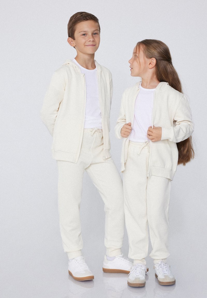 Weiße Baumwoll-Zip-Hoodies mit passenden Jogginghosen. Glatte Textur, lockere Passform und elastische Bündchen. Kombiniert mit weißen Sneakers, Seite an Seite stehend.