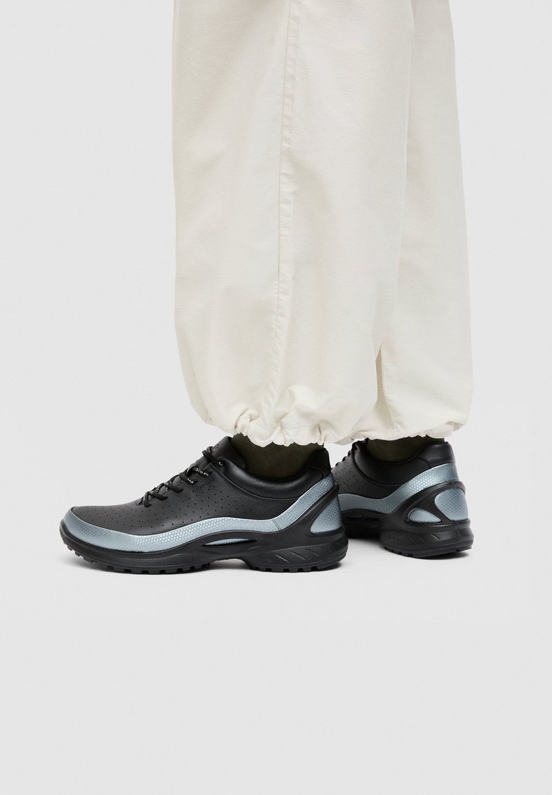 Chaussures de sport à lacets noires et grises portées avec un pantalon beige à bords élastiques, reposant sur un sol gris clair.