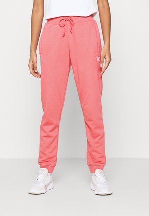 Personne portant un pantalon de survêtement Adidas rose corail avec taille à cordon et des baskets blanches, debout devant un fond clair uni.