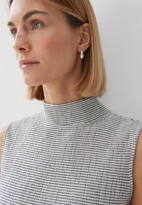 Kraagloze, mouwloze top in zwart-wit ruitpatroon; gladde textuur; zilveren druppeloorbellen; natuurlijk, steil haar.