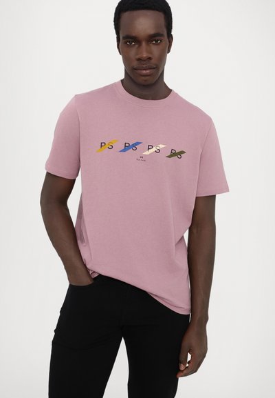 Růžové bavlněné tričko s krátkým rukávem, které má na hrudi vícolorovaný design loga 'PS'. Model má na sobě černé kalhoty.