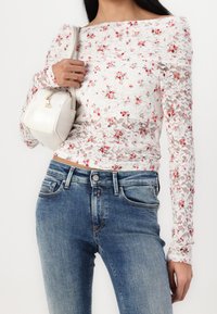 Mujer con una blusa blanca de encaje floral de manga larga y jeans azules, llevando un pequeño bolso blanco al hombro frente a un fondo liso.