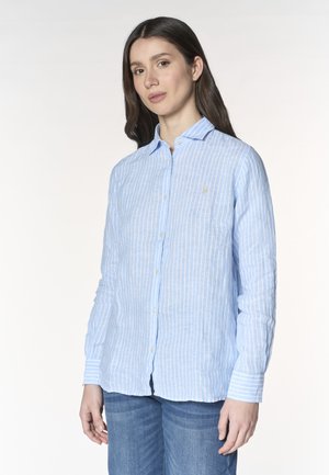 Donna con capelli lunghi e scuri che indossa una camicia a righe azzurro chiaro e bianco con bottoni e jeans blu su uno sfondo semplice.