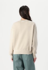Felpa beige dal taglio casual, con polsini e orlo a costine. Dettaglio di cucitura abbinato. Indossata con pantaloni verdi larghi.