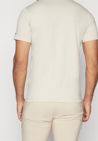 Mand iført en lys beige kortærmet t-shirt og matchende beige bukser, vist fra ryggen mod en ensfarvet baggrund.