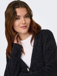 Noorpruunide pikade juustega noor naine kannab musta karvast kardigaani valge särke peal, hellalt kaela puudutades, ühevärvilise tausta ees.