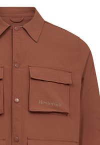 Camisa marrón de botones fabricada en un tejido suave, con dos bolsillos frontales y el logo "Resteröds" en gris claro en el bolsillo izquierdo.