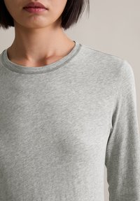 Helles graues Langarmshirt aus einem weichen, strukturierten Stoff. Es hat einen runden Ausschnitt und subtile Stickdetails.