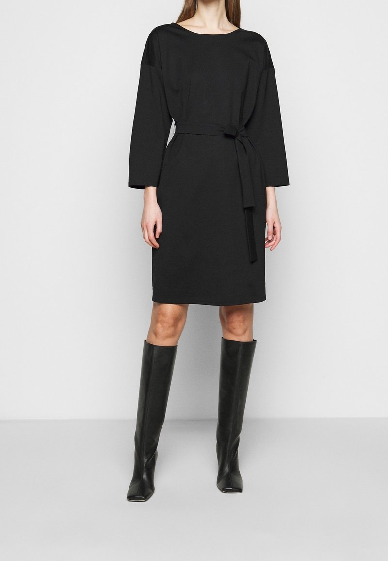 Femme portant une robe noire longueur genou avec manches longues et ceinture à la taille, associée à des bottes noires montant jusqu'aux genoux.