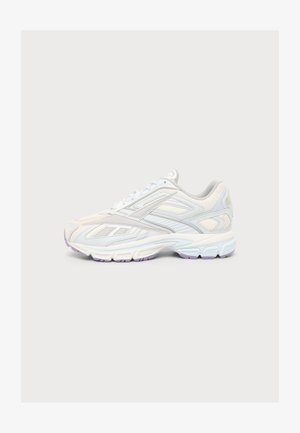 Zapatilla deportiva blanca y gris claro con suela texturizada y diseño con cordones, mostrada de lado sobre un fondo claro y liso.