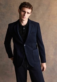 Marinblå ullblazer med slag, två framfickor och stängning med en knapp, kombinerad med en svart polo och mörk denimjeans.