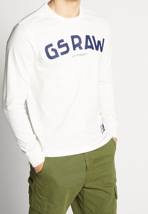 Hombre que lleva una camisa blanca de manga larga con el texto "GS RAW" en el pecho y pantalones cargo verde oliva, con las manos en los bolsillos.