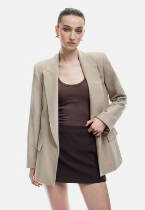 Beige Blazer mit strukturierten Schultern, breiten Revers und zwei vorderen Taschen, getragen über einem braunen gerippten Tanktop und einem braunen Minirock.