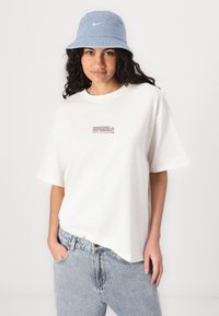 Camiseta de algodón blanca con el logo impreso de "Ripcurl" en rojo, combinada con unos pantalones vaqueros de mezclilla azul claro y un sombrero tipo bucket de color azul claro a juego.