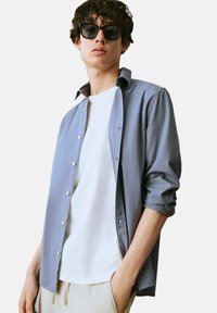 Camicia button-up grigia chiara con maniche arrotolate, indossata sopra una maglietta bianca. Il modello ha i capelli corti e ricci e occhiali da sole neri.