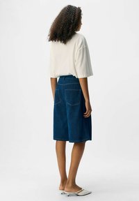 Donna con capelli ricci che indossa una camicia bianca a maniche corte larga, pantaloni di jeans corti fino al ginocchio e scarpe basse bianche senza lacci, in posizione laterale.